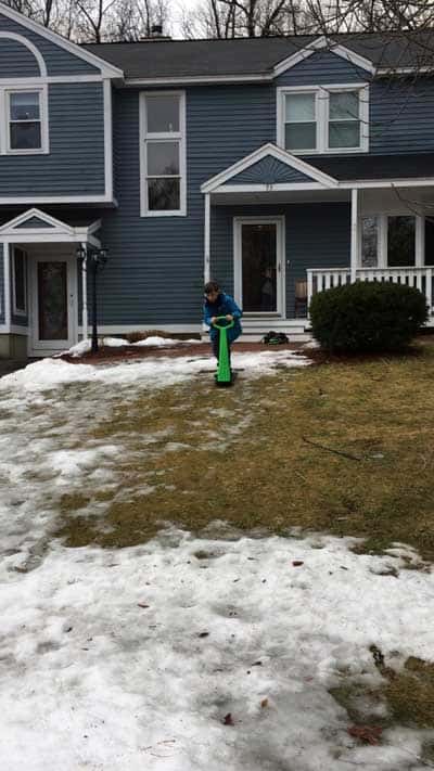 A boy on a snow scooter on the wet grass with a little snow in front of his home 