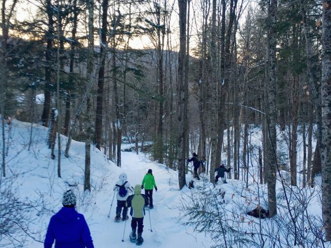 6 people of varying ages hiking through the woods on snowshoes on a wooded snowy trail