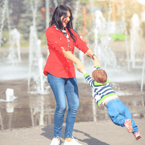 mom swinging her child by the arms near a water fountain having fun in the summer 