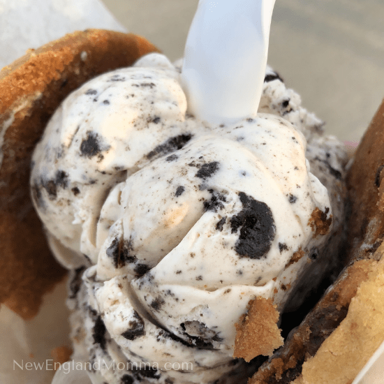 a scoop of cookies and cream ice cream sandwiched between two soft looking chocolate chip cookies and a spoon. 
