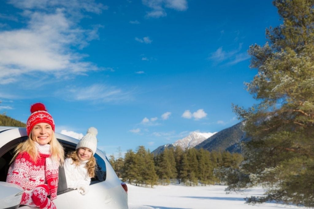 Mom and daughter with winter sweaters and hats, looking out a car to a field of snow and trees