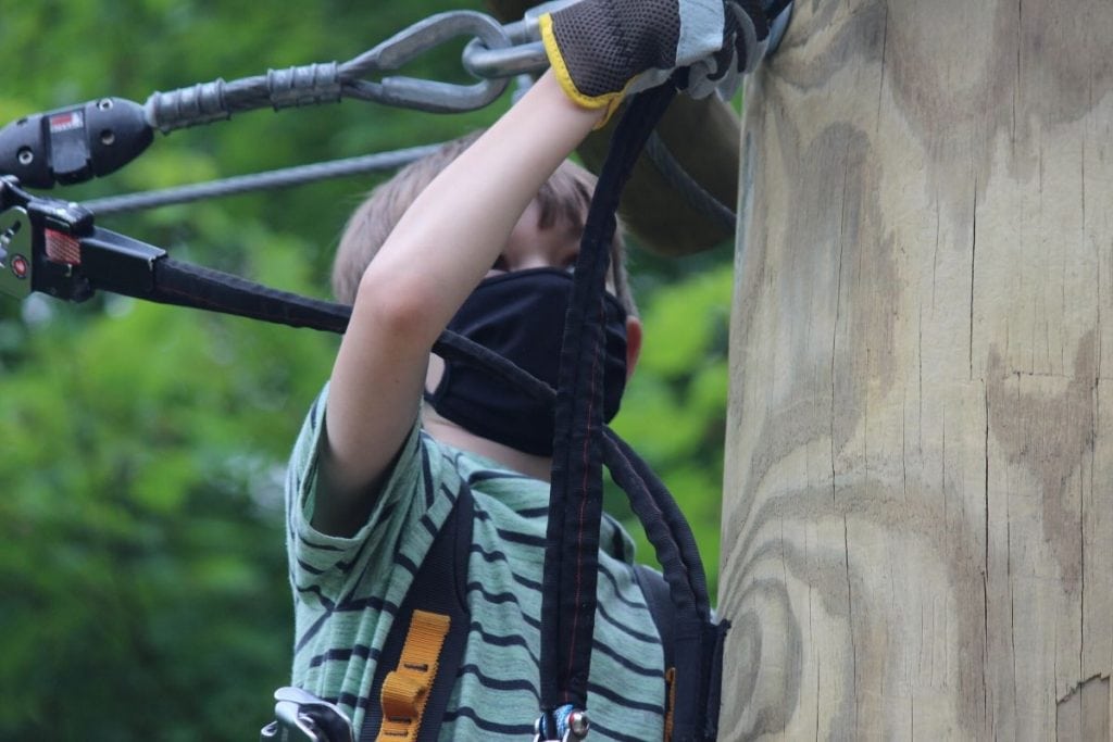 A kid with a mask on being socially distant while having fun out on a rope course outdoors at Boundless. 