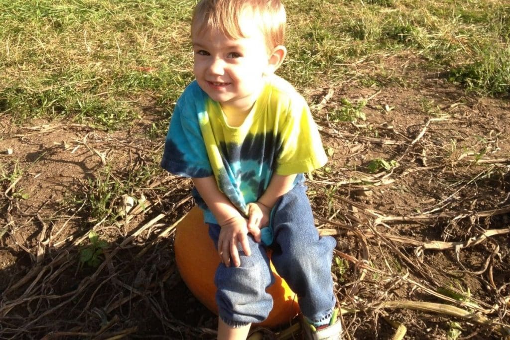 A young boy sitting on an orange pumpkin in a hay field