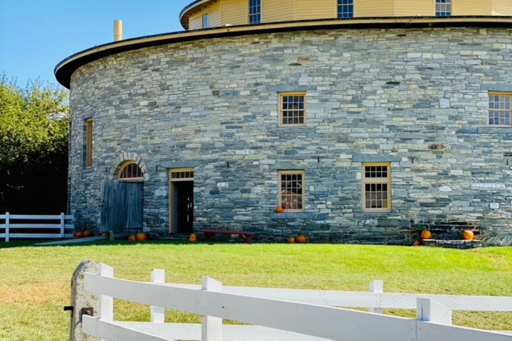 A round stone barn with a white picket fence and pumpkins outdoors for the fall great to visit for a weekend in the Berkshires